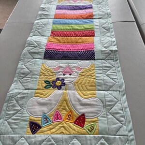 May include: A quilted Easter table runner featuring a bunny design with colorful accents. The runner has a light blue border and a central section with a rainbow of colors and the word "EASTER". The bunny is white with yellow and pink details.