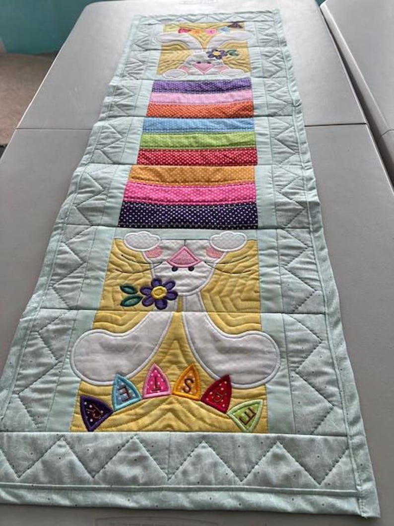May include: A quilted Easter table runner featuring a bunny design with colorful accents. The runner has a light blue border and a central section with a rainbow of colors and the word "EASTER". The bunny is white with yellow and pink details.