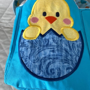 May include: A handmade, turquoise blue tote bag with a whimsical chick design. The chick is yellow with an orange beak and pink cheeks, emerging from a blue patterned egg. The bag has a matching patterned handle.