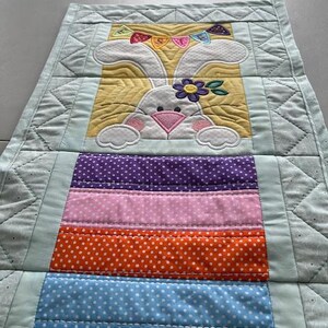 May include: A quilted Easter table runner featuring a white bunny with a flower and a banner. The runner has a yellow background and colorful polka-dot stripes in purple, pink, orange, blue, and green. The edges are light blue.