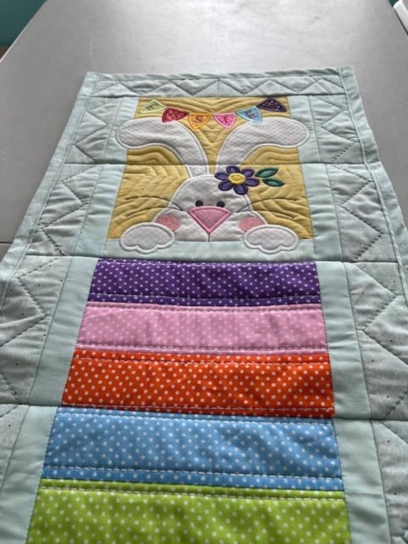 May include: A quilted Easter table runner featuring a white bunny with a flower and a banner. The runner has a yellow background and colorful polka-dot stripes in purple, pink, orange, blue, and green. The edges are light blue.