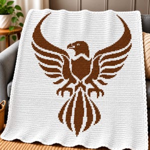 Könnte beinhalten: Eine weiße Häkeldecke mit einem großen, zentrierten braunen Adler-Motiv mit ausgebreiteten Flügeln. Die Decke hat eine texturierte, gestrickte Oberfläche und einen gewellten Rand, der einen klaren Kontrast zwischen braunem und weißem Garn zeigt.