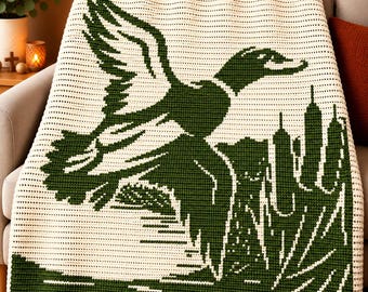 May include: A cream-colored crochet blanket featuring a large, dark green graphic of a duck in flight over a marshy landscape with tall reeds. The design is created using a repeating stitch pattern, showcasing a high-contrast two-tone color scheme.