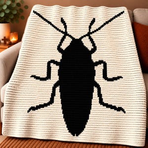 May include: A cream-colored crochet blanket featuring a large, centered black insect silhouette. The blanket is constructed with a horizontal stitch pattern, displaying a clear contrast between the dark bug graphic and the light background.