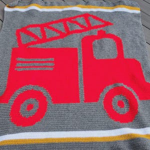 May include: A gray crocheted blanket featuring a large red fire truck graphic with a ladder on top. The blanket has horizontal white and yellow stripes along the top and bottom edges, creating a road-like design with a textured yarn finish.