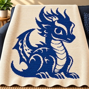 May include: A cream-coloured crochet blanket featuring a large, centred graphic of a blue dragon. The dragon is depicted in a seated position with its wings partially spread, showcasing a detailed, textured stitch pattern throughout the fabric.