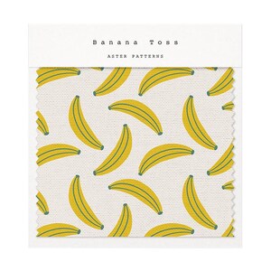Puede incluir: Muestra de tela con un patr&oacute;n repetido de pl&aacute;tanos amarillos con detalles verdes sobre un fondo crema. El texto "Banana Toss" y "Aster Patterns" est&aacute; impreso en la parte superior. La tela tiene una apariencia texturizada.