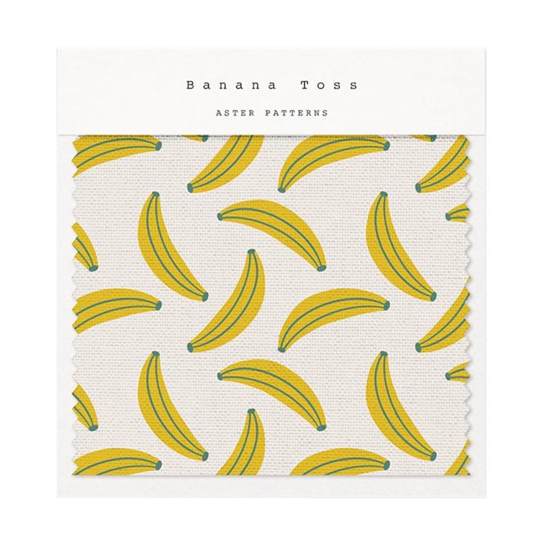 Puede incluir: Muestra de tela con un patr&oacute;n repetido de pl&aacute;tanos amarillos con detalles verdes sobre un fondo crema. El texto "Banana Toss" y "Aster Patterns" est&aacute; impreso en la parte superior. La tela tiene una apariencia texturizada.