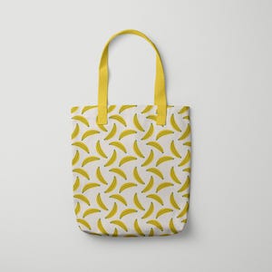 Puede incluir: Un bolso tote blanco con un patr&oacute;n repetido de pl&aacute;tanos amarillos. El bolso tiene asas amarillas y probablemente est&aacute; hecho de tela. El dise&ntilde;o es simple y llamativo, adecuado para llevar art&iacute;culos personales.