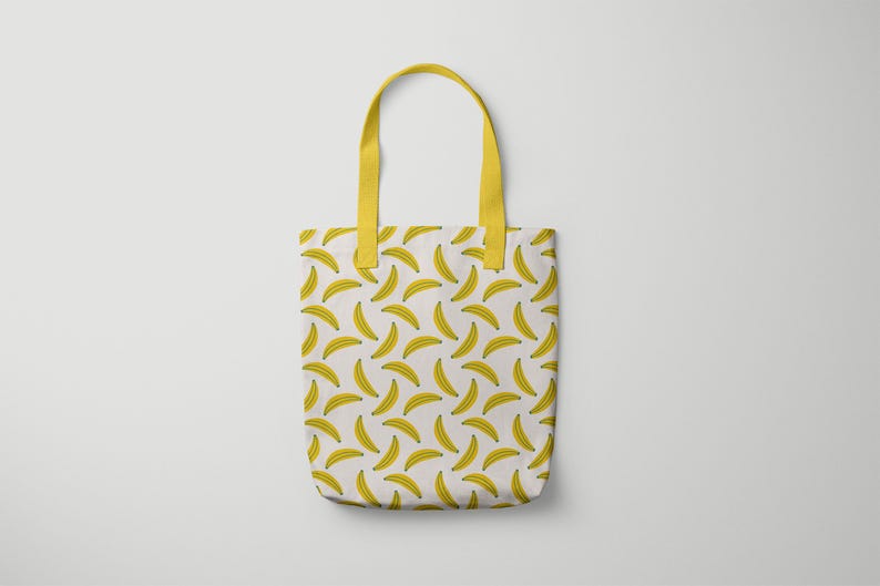 Puede incluir: Un bolso tote blanco con un patr&oacute;n repetido de pl&aacute;tanos amarillos. El bolso tiene asas amarillas y probablemente est&aacute; hecho de tela. El dise&ntilde;o es simple y llamativo, adecuado para llevar art&iacute;culos personales.