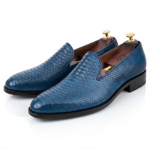 May include: A pair of blue slip-on loafers featuring a textured snakeskin pattern, a rounded toe, and a black sole. Each shoe includes a wooden shoe tree with a gold-toned metal knob inserted inside to maintain the shape of the footwear.