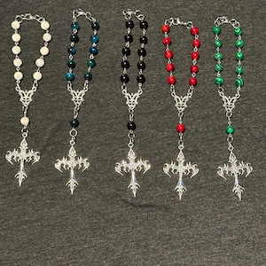May include: Five beaded rosaries with silver-colored crosses. The beads are white, teal, black, red, and green. The crosses have an ornate design. The rosaries are displayed on a gray surface.