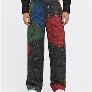 May include: Full-length trousers featuring a multi-colored patchwork design in shades of red, blue, green, and black. The fabric is covered in a repeating print of metallic silver safety pins scattered across the surface of the pants.