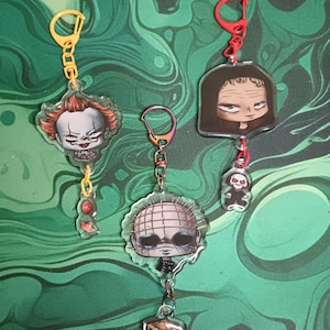 Könnte beinhalten: Drei Acryl-Schlüsselanhänger mit Horrorfiguren. Einer hat einen gelben Verschluss mit einem Clown und einem roten Ballon. Ein weiterer hat einen schillernden Verschluss mit einer Figur mit Nadelkopf und einer Puzzlebox. Der dritte hat einen roten Verschluss.