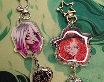 Puede incluir: Dos llaveros acrílicos con ilustraciones de dibujos animados. Uno presenta a una mujer con cabello blanco y rosa y un dije de taza de café. El otro tiene una mujer con cabello rojo, gafas y un dije de libro. Ambos tienen cierres metálicos.