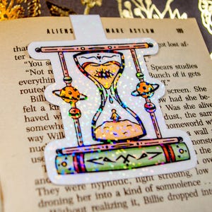 May include: A holographic sticker featuring an hourglass with a celestial design. The hourglass is filled with glitter and has a book with the word "Camp" on it. The sticker is on a book with the text "Aliens Make Asylum 105".