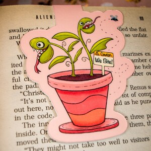 Puede incluir: Una ilustración de dibujos animados rosa y verde de una planta carnívora con un cartel de advertencia que dice "¡Cuidado, mordemos!" La planta está en una maceta rosa con tierra marrón y tiene una boca con dientes.