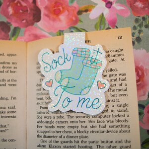 以下が含まれることがあります： キラキラ光るしおりに、薄いブルーの靴下と「Sock it to me」の文字が書かれています。靴下はピンクと白のチェック柄で、ピンクのハートが付いています。