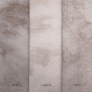 May include: Three vertical fabric swatches in shades of taupe and beige. The left swatch is Aida cloth with a visible grid, the center is smooth Lugana fabric, and the right is textured Linen. Each piece features a mottled, hand-dyed appearance.