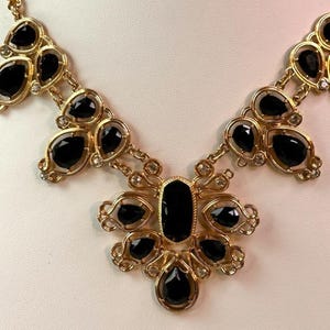Puede incluir: Un collar dorado con un grupo central de piedras negras en forma de lágrima y una gran piedra negra rectangular. El collar tiene una cadena y grupos más pequeños de piedras negras con detalles transparentes.