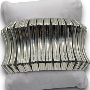 May include: A silver-toned bracelet with a ribbed design, resting on a white pillow. The bracelet has a curved shape and is made of multiple vertical bars. The jewelry is displayed against a white background.