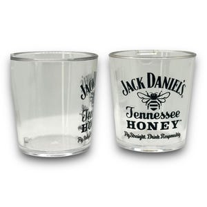 May include: Two clear plastic shot glasses featuring the black printed Jack Daniel's Tennessee Honey logo with a bee graphic. The text reads Jack Daniel's Tennessee Honey and Fly Straight. Drink Responsibly. The glasses have a cylindrical shape.