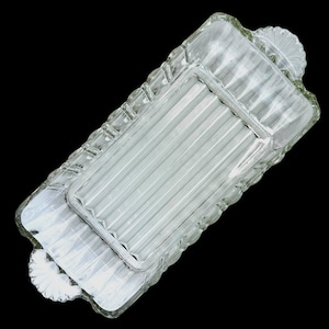 May include: Two clear glass serving trays. The larger tray has a rectangular shape with a ridged pattern and scalloped edges. The smaller tray fits inside the larger one and has a similar ridged design. Both trays have decorative handles.