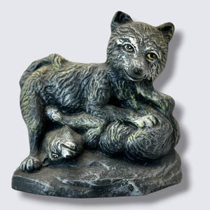 May include: A gray and black wolf figurine featuring two wolves interacting on a textured rock base. The standing wolf has detailed fur texture and yellow eyes, while the second wolf is positioned underneath, both rendered in a stone-like finish.