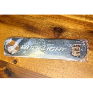 May include: A silver-colored bottle opener with the "BUD LIGHT" logo in white lettering. The opener is rectangular with a circular opening on one end and is packaged in clear plastic.