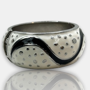 May include: A wide, silver-toned bangle bracelet with a cream-colored enamel finish. The bracelet features a black wavy design and small, silver-colored dots. The bracelet is reflected in the surface below.