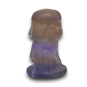 May include: A carved figurine of a character with a rounded head and flowing robes. The figurine is made of a translucent stone with shades of purple, brown, and white. The base is a darker shade of purple.