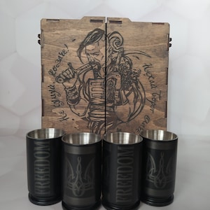 Puede incluir: Una caja de madera con tapa con bisagras, que presenta una imagen grabada con láser de un hombre con barba y una espada. La caja contiene cuatro vasos de chupito negros con la palabra "FREEDOM" y un símbolo de tridente grabado en ellos. La caja está etiquetada como "¡Gloria a los héroes!"