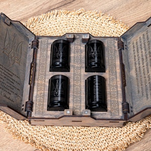 Op de afbeelding: Een houten doos met een scharnierend deksel, gegraveerd met een Oekraïens drietandsymbool en een citaat. De doos bevat vier zwarte shotglazen, die ook allemaal gegraveerd zijn met het drietandsymbool.