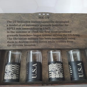Puede incluir: Un juego de caja de madera con cuatro vasos de chupito transparentes. Las bases negras llevan "U.S.A." y motivos de balas. La caja tiene texto grabado, incluyendo "MK-19" e información sobre un lanzagranadas y el ejército ucraniano.