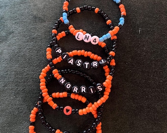May include: A collection of beaded bracelets in black, orange, and blue. Each bracelet features white letter beads spelling out names and initials, with heart-shaped beads. The bracelets are on a dark green surface.