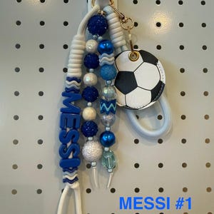 May include: A keychain featuring a white cord, blue beads, and a round soccer ball charm. The blue beads spell out the name MESSI. The design includes glittery blue, white, and translucent beads attached to a gold-toned metal ring and clasp.