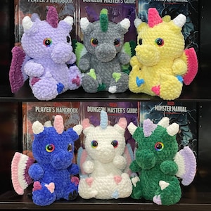 May include: Six crocheted dragon plushies in different colors, including purple, gray, yellow, blue, white, and green. Each plush has a horn, wings, and rainbow eyes. The plushies are arranged in two rows of three, with the top row sitting on top of three books titled "Player's Handbook", "Dungeon Master's Guide", and "Monster Manual".