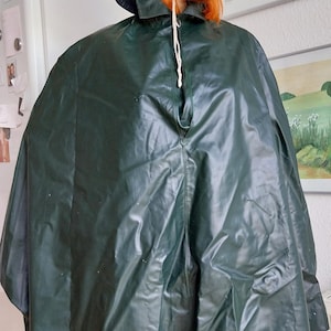 May include: A dark green rain poncho with a matching hood. The poncho has a drawstring closure and a pocket on the right side. The mannequin is wearing a black hood with a drawstring.