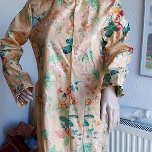May include: A long, peach-colored raincoat with a floral pattern of red, green, and white flowers and leaves. The coat is buttoned up and has a collar. The mannequin is wearing a red wig.