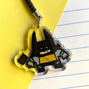 Puede incluir: Un charm de acrílico transparente con un diseño de Batman de dibujos animados. El charm tiene un cordón negro y un cierre de metal plateado. Batman está representado en negro, gris y amarillo, con un diseño estilizado y un pequeño accesorio en forma de murciélago.