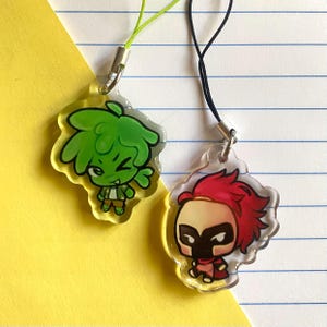 May include: Two clear acrylic charms with cartoon character designs. One charm features a green character with spiky hair and a mischievous expression. The other charm has a red-haired character wearing a black mask. Each charm has a colored lanyard.