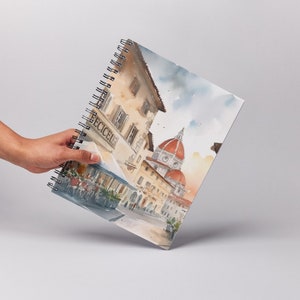 K&ouml;nnte beinhalten: Ein spiralgebundenes Notizbuch mit einer Aquarell-Illustration einer Stra&szlig;enszene in Florenz, Italien. Die Illustration zeigt ein Caf&eacute; mit einem Schild, auf dem "ECCEICI" steht, und den ber&uuml;hmten Dom im Hintergrund.
