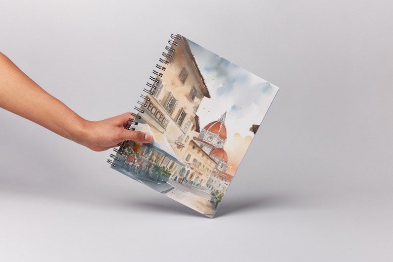 K&ouml;nnte beinhalten: Ein spiralgebundenes Notizbuch mit einer Aquarell-Illustration einer Stra&szlig;enszene in Florenz, Italien. Die Illustration zeigt ein Caf&eacute; mit einem Schild, auf dem "ECCEICI" steht, und den ber&uuml;hmten Dom im Hintergrund.