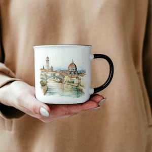 K&ouml;nnte beinhalten: Wei&szlig;e Keramiktasse mit schwarzem Henkel. Die Tasse zeigt eine Aquarell-Illustration einer Stadtlandschaft mit einer Br&uuml;cke und einem kuppelf&ouml;rmigen Geb&auml;ude.