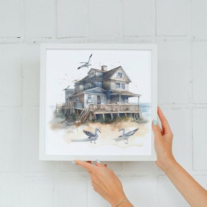 May include: A watercolor painting of a blue house on stilts with a white picket fence and a porch. The house is surrounded by sand and seagulls. The painting is framed in a white frame.