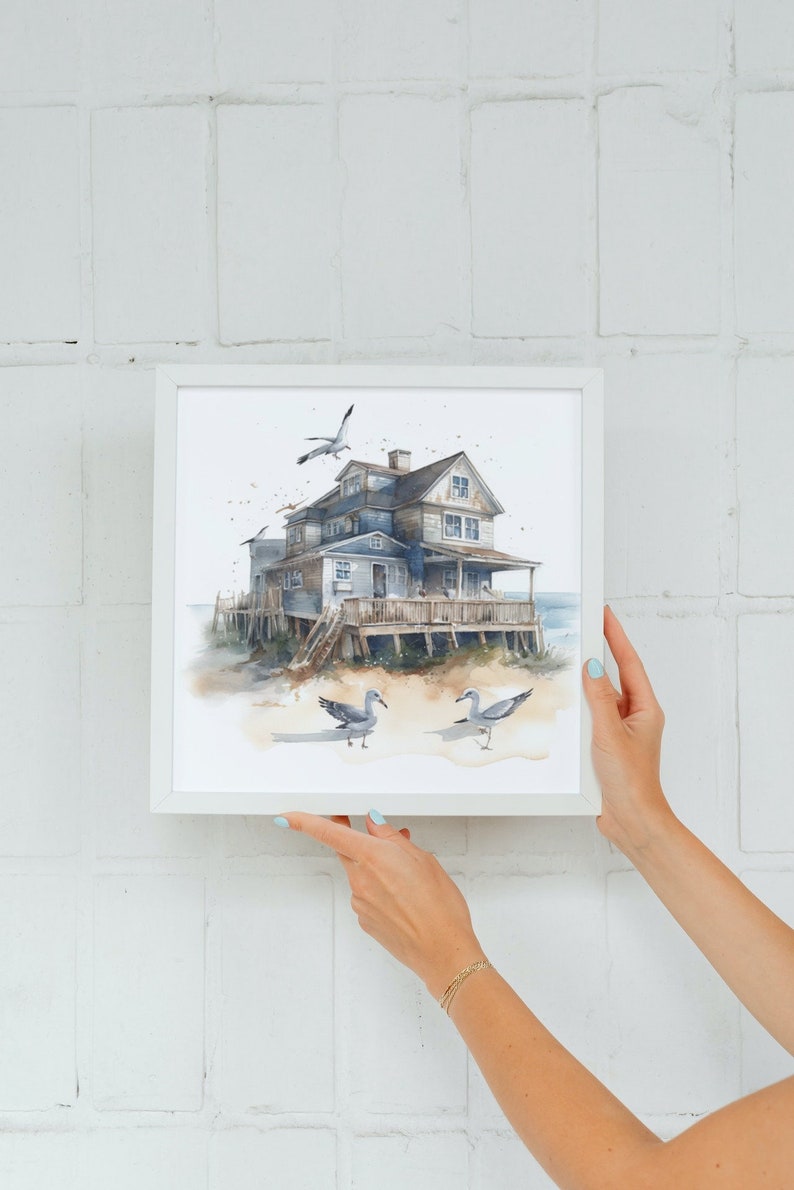May include: A watercolor painting of a blue house on stilts with a white picket fence and a porch. The house is surrounded by sand and seagulls. The painting is framed in a white frame.