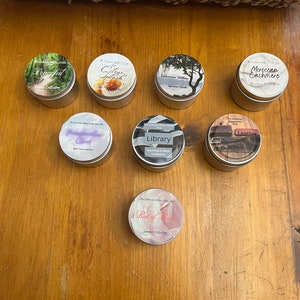 Könnte beinhalten: Sortiment von acht kleinen, runden, silbernen Kerzenbehältern mit verschiedenen naturinspirierten Designs und Etiketten. Die Etiketten beinhalten Namen wie "Peaceful Sunset", "Citrus Splash", "Bathhouse Willow", "Moroccan Cashmere", "Dreamweaver", "Library", "Leather" und "Bed of Roses".