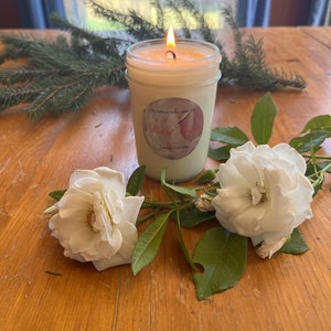 Può includere: Una candela bianca in un barattolo di vetro trasparente con un'etichetta che recita "The Catherine Willow Candle Co. 2023" e "Bed of Roses" in lettere rosa. La candela è circondata da rose bianche e foglie verdi su una superficie di legno.