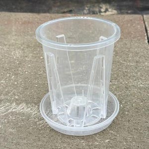 May include: A clear, cylindrical plastic plant pot featuring a raised internal structure with vertical supports and a central drainage cone, accompanied by a matching circular saucer base designed for drainage and airflow for plant roots.