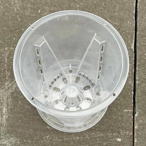 May include: A clear plastic plant pot featuring a raised central cone and vertical side supports with integrated drainage slots along the base and walls, designed for optimal airflow and water drainage for potted plants.